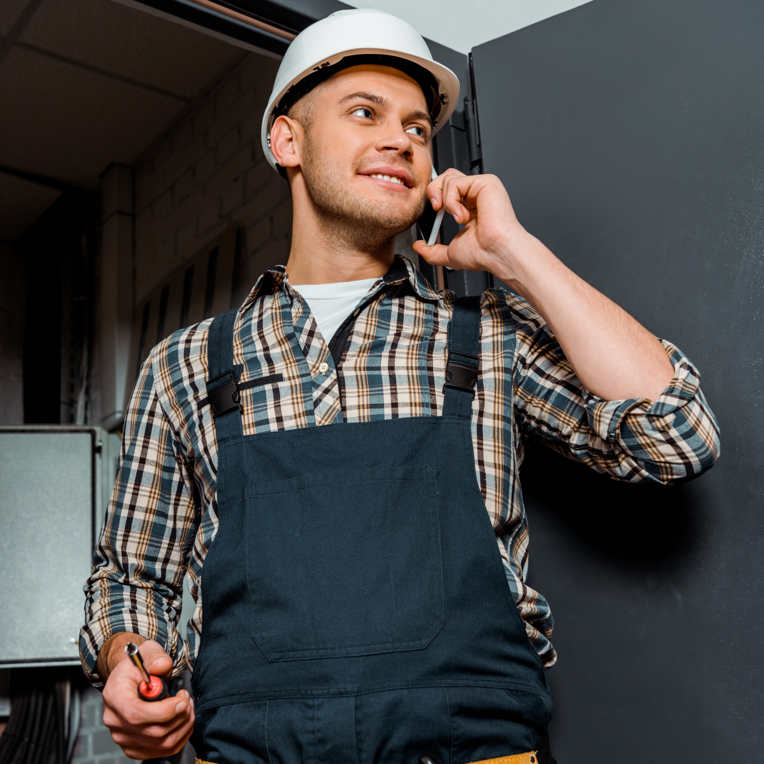 cheerful installer in safety helmet holding screwdriver while talking on smartphone Fröhlicher Installateur mit Schutzhelm, der einen Schraubenzieher in der Hand hält, während er mit seinem Smartphone telefoniert.