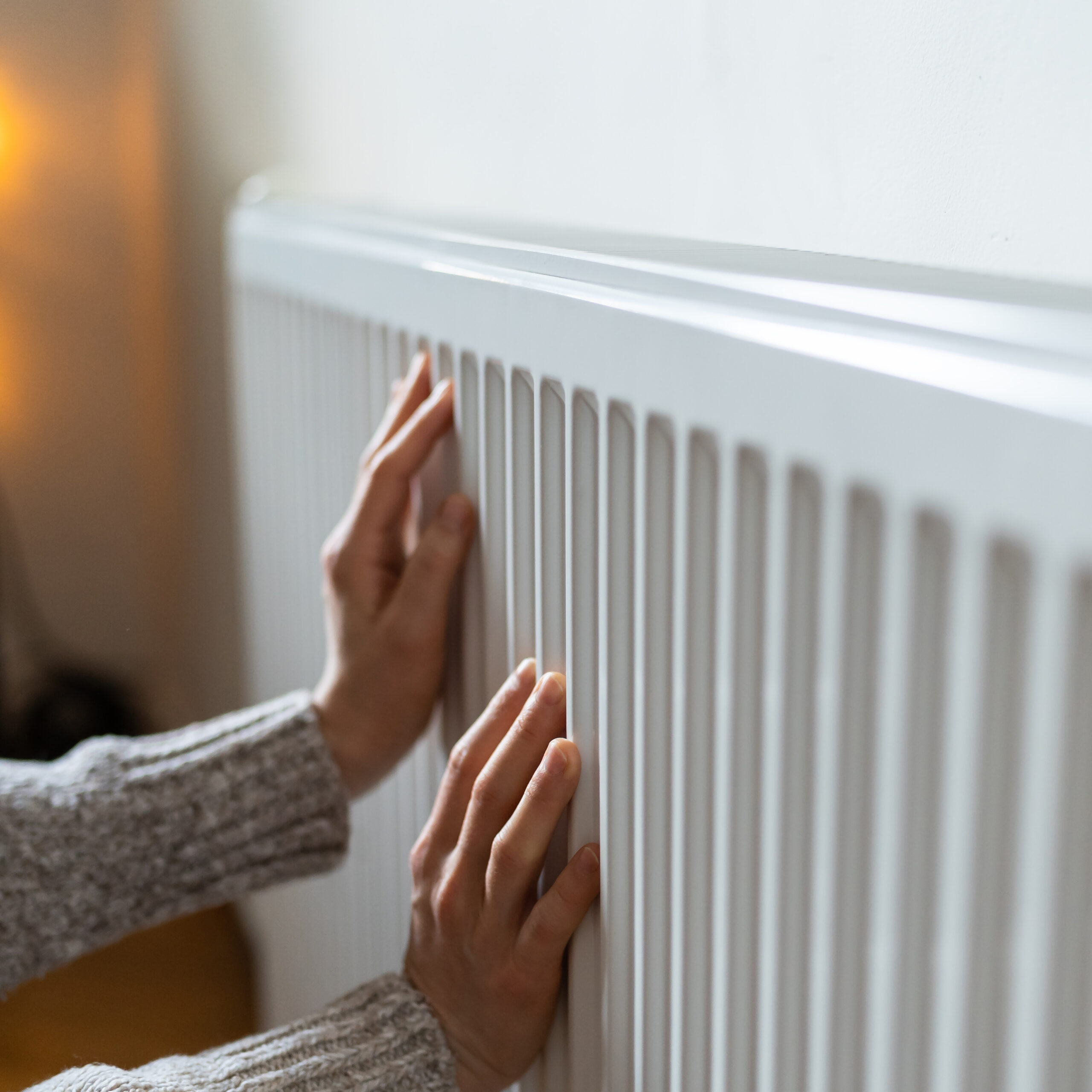 Closeup of woman in sweater puts hands on room central heating battery to warm up in cold apartment Nahaufnahme einer Frau in einem Wollpullover, die die Wärme in ihrer Wohnung bewahren möchte. Die Frau legt ihre Hände auf die Zentralheizungsbatterie im Zimmer, um sich aufzuwärmen und Krankheiten aufgrund der Kälte vorzubeugen. Die Person überprüft die Funktion der Heizung.
